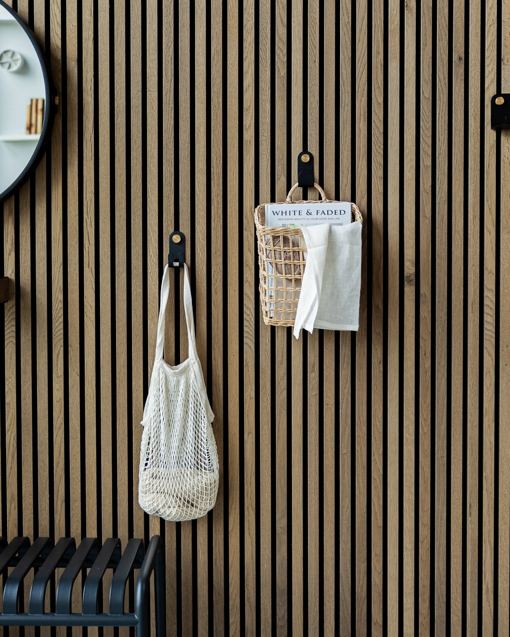 Una bolsa de malla y una cesta cuelgan de los Single Hangers Norr de WoodUpp sobre un Panel acústico Akupanel de madera nórdica; se ve parte de un espejo a la izquierda.